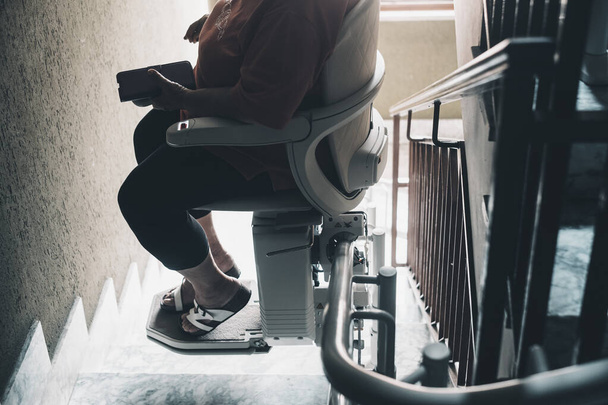 Пожилой женщине нужна современная помощь, чтобы подняться домой. Концепция возрастных проблем. Электрический стул для людей с ограниченными возможностями на ковровой лестнице в жилом доме. - Фото, изображение