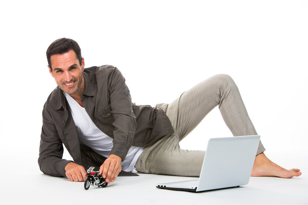 Mies makaa lattialla hymyillen kameralle leikkien moottoripyörän pienoismallilla ja kannettavalla hänen vieressään
 - Valokuva, kuva
