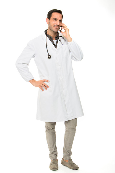 Volledige lengte portret os een lachende mannelijke arts met een stethoscoop tijdens het gebruik van een mobiele telefoon - Foto, afbeelding