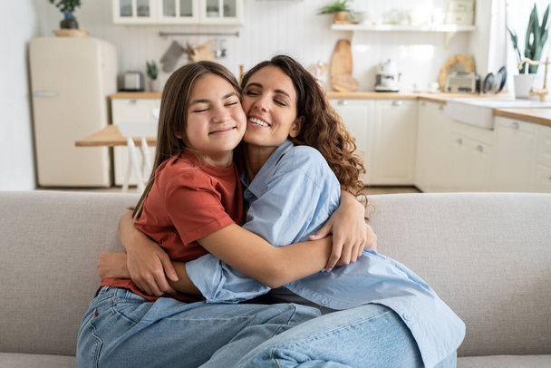 Happiness in motherhood. Joyful young mother and teen daughter cuddling hugging while sitting on sofa together, excited overjoyed girl child embracing mom coming back home. Family wellbeing concept - Fotó, kép