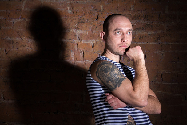 Серьезный лысый мужчина с татуировкой, стоящий у кирпичной стены
 - Фото, изображение