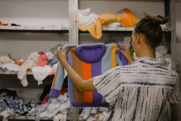 Молодая женщина с разноцветной пурпурной полосатой шерстью вязала детский свитер. Человек выбирает теплую одежду для ребенка в пункте распределения гуманитарной помощи для беженцев и обездоленных. - Фото, изображение