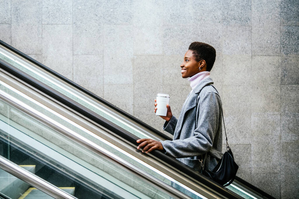 Side view of stylish African American female in stylish coat and sweater. She is with cup of coffee and bag smiling and riding moving stairs in city - Foto, Bild