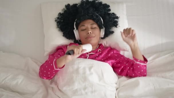 Funny adorable happy African American girl actively dancing in bed, singing in sticky roller microphone. Cute woman of color having fun dancing to music playing in bedroom alone. Enjoy singer morning - Filmmaterial, Video
