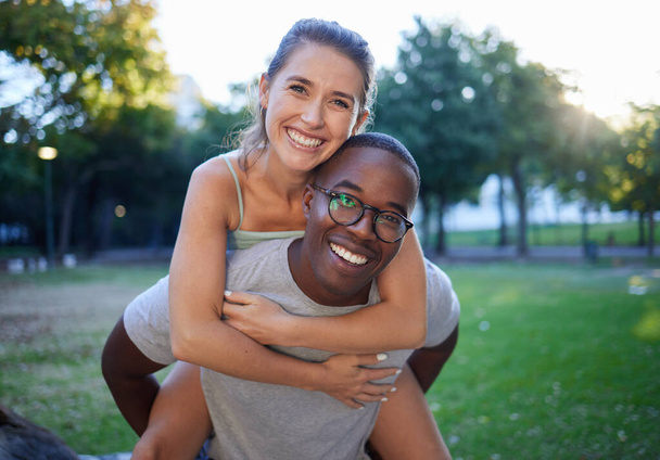 Portrait, love and piggyback with an interracial couple together outdoor in a park for bonding or a date. Summer, diversity and smile with a man and woman happy outside in nature while dating. - Photo, image