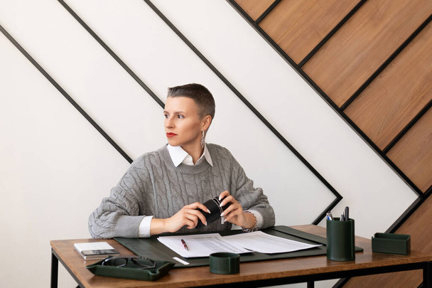 Деловая женщина с короткой стрижкой смотрит в окно, сидя за столом в офисе. - Фото, изображение