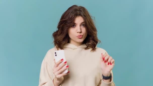 Portrait of Happy cute gorgeous and happy brunette latin woman listening to music on mobile smartphone singing and dancing carefree on blue background. copy space - Video
