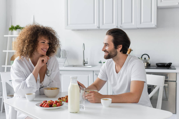 cheerful young woman having breakfast with bearded boyfriend in kitchen  - Foto, Imagen