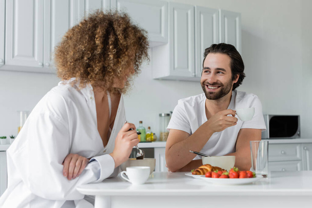 веселый бородатый мужчина улыбается, глядя на кудрявую подружку во время завтрака  - Фото, изображение
