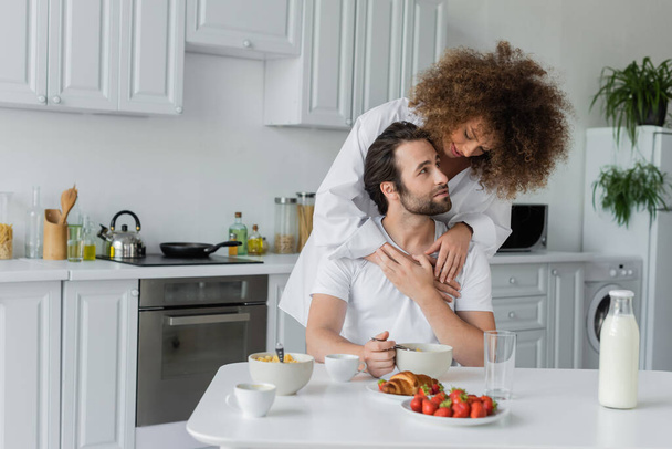 curly young woman hugging bearded boyfriend during breakfast in kitchen  - Fotografie, Obrázek