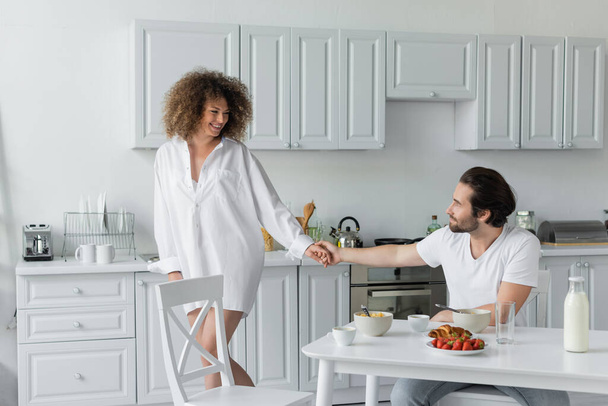 бородатый мужчина, держащийся за руки с веселой женщиной во время завтрака на кухне  - Фото, изображение