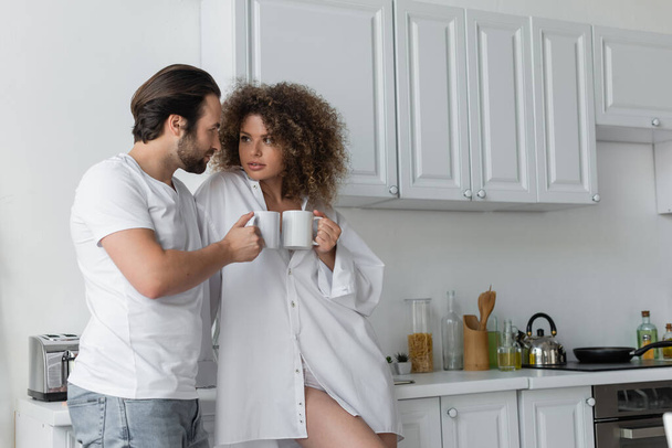 bearded man and curly woman holding cups of coffee in kitchen  - Photo, image