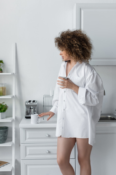 curly young woman in white shirt standing with cup of coffee in kitchen  - Fotó, kép