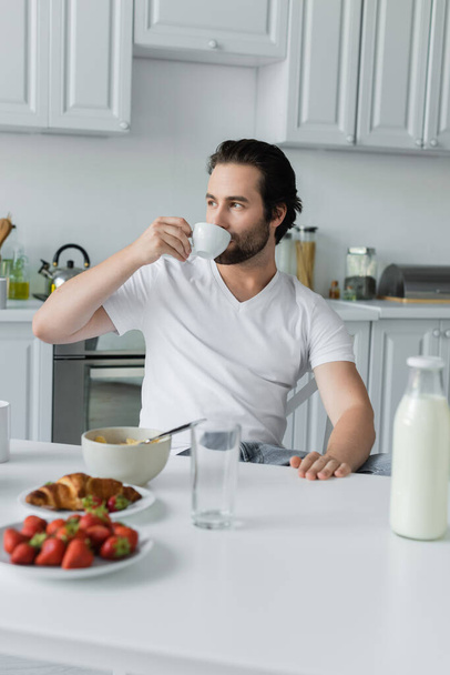 bearded man drinking coffee near tasty breakfast on table in kitchen  - Foto, Bild
