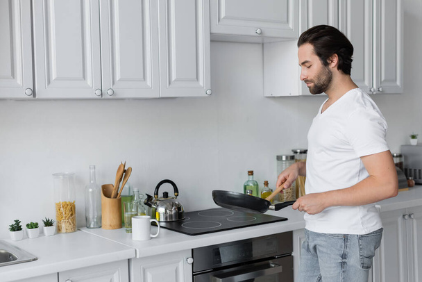 бородатый мужчина в джинсах и белой футболке готовит завтрак на кухне  - Фото, изображение