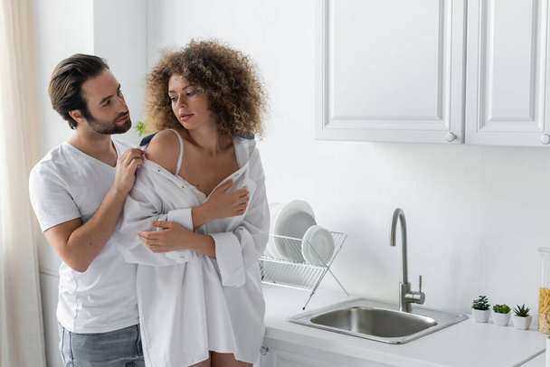 bearded man undressing curly woman in white shirt in kitchen  - Foto, afbeelding