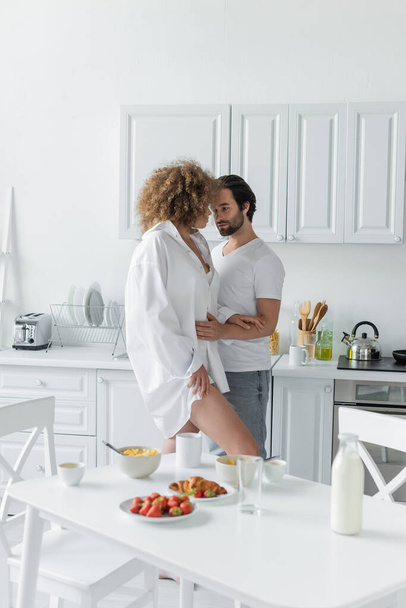 Молодой человек в джинсах соблазняет чувственную женщину в белой рубашке на кухне  - Фото, изображение