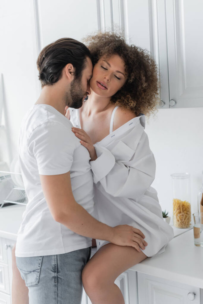 bearded man in jeans seducing curly young woman with closed eyes sitting on kitchen worktop  - Foto, Bild
