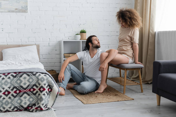 bearded man sitting on carpet and looking at curly girlfriend on bed bench  - Φωτογραφία, εικόνα