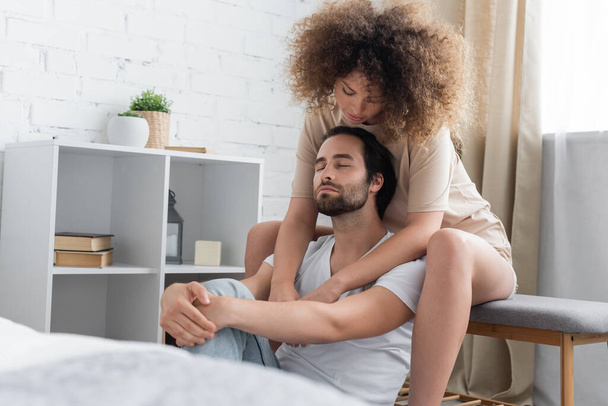 curly young woman sitting on bed bench hugging bearded boyfriend with closed eyes in bedroom  - Foto, immagini