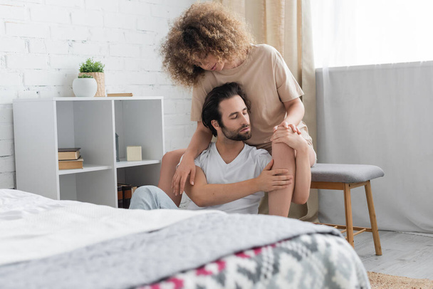 curly woman sitting on bed bench hugging bearded boyfriend with closed eyes in bedroom  - Фото, зображення