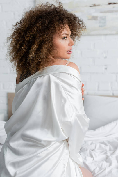 side view of curly and sexy young woman in white silk robe sitting on bed  - Foto, Imagen