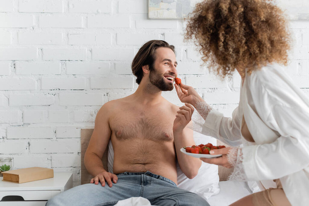curly woman feeding cheerful boyfriend with ripe strawberry  - Foto, afbeelding