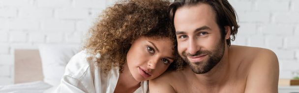 curly woman in silk robe leaning on shirtless boyfriend smiling in bedroom, banner  - Photo, image