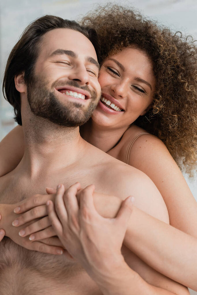 cheerful and curly young woman hugging shirtless man at home  - Photo, image