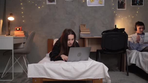 young teenager girl student lies on a bed in a college dormitory at night looking for information online using her laptop. copy space. - Filmati, video
