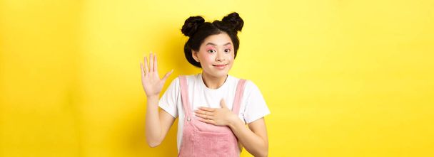 Obraz uroczej azjatyckiej dziewczyny z makijażem jest uczciwy, podnosząc rękę i umieścić na sercu, mówiąc prawdę, dać przysięgę i uśmiech, stojąc na żółtym tle. - Zdjęcie, obraz