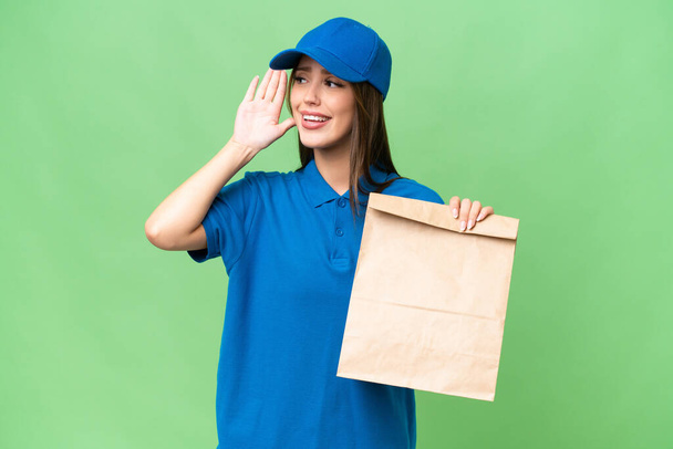 Young beautiful caucasian woman taking a bag of takeaway food over isolated background listening to something by putting hand on the ear - Фото, изображение