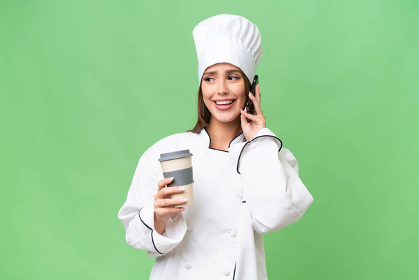 Jonge chef-kok Kaukasische vrouw over geïsoleerde achtergrond houden koffie mee te nemen en een mobiele - Foto, afbeelding