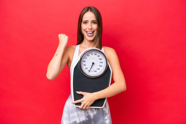Young beautiful caucasian woman isolated on red background with weighing machine and doing victory gesture - Valokuva, kuva