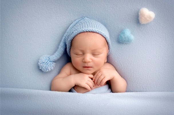 A cute newborn boy in the first days of life sleeps naked on a blue fabric background in a blue knitted wool cap. Light blue and pink felt heart on a blue background.  - Foto, Bild