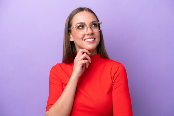 Young caucasian woman isolated on purple background With glasses and thinking while looking up - Фото, изображение