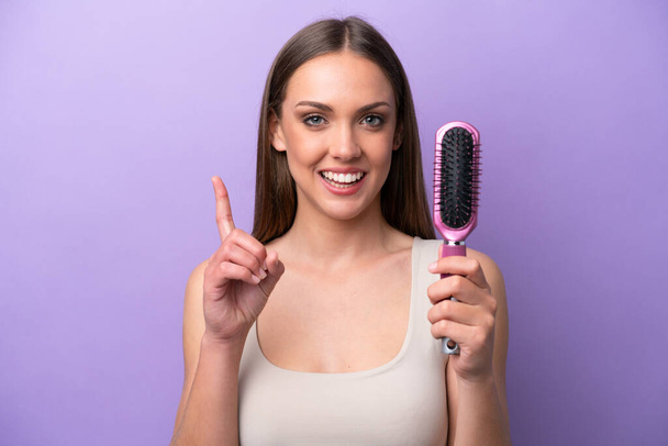 Young caucasian woman holding hairbrush isolated on purple background pointing up a great idea - 写真・画像