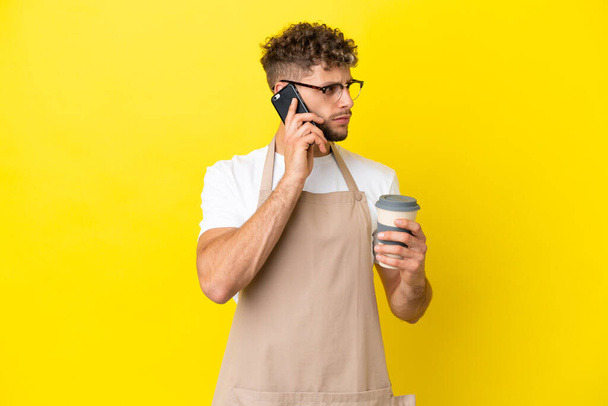 Офіціант ресторану блондинка ізольована на жовтому фоні, тримаючи каву, щоб забрати і мобільний
 - Фото, зображення