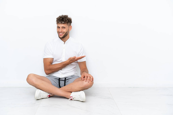 Young blonde man sitting on the floor isolated on white background presenting an idea while looking smiling towards - Fotoğraf, Görsel