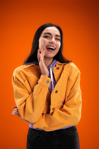 Excited brunette woman in jacket and hoodie looking at camera isolated on orange - Foto, imagen