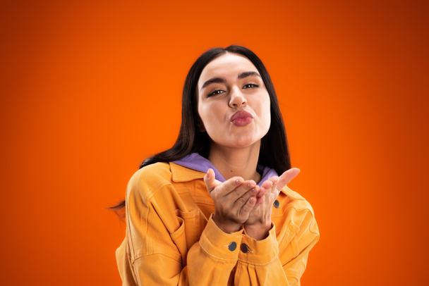 Nuori brunette nainen puhaltaa ilmaa suudella kameran eristetty oranssi - Valokuva, kuva
