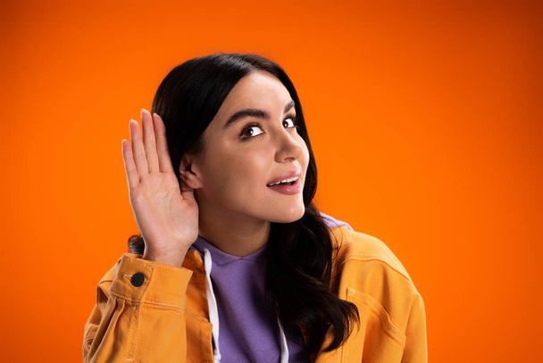 Молодая женщина в куртке слушает, держа руку возле уха изолированы на оранжевый - Фото, изображение