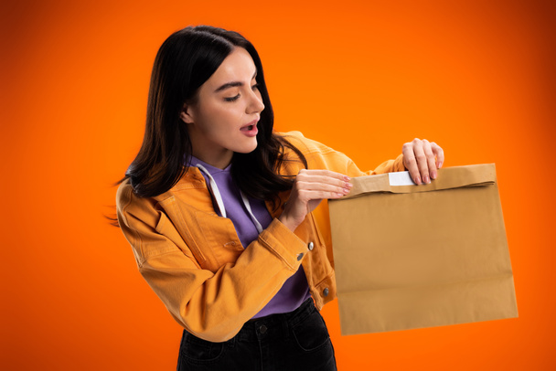 Excited brunette woman looking at paper bag isolated on orange - Fotografie, Obrázek