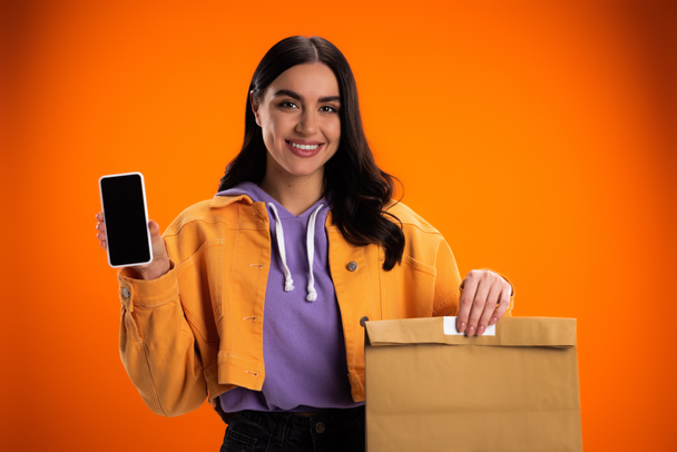 Smiling woman holding paper bag and smartphone with blank screen isolated on orange - Zdjęcie, obraz
