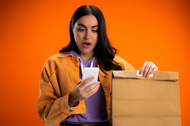 Shocked woman using smartphone and holding takeaway paper bag isolated on orange - Photo, Image