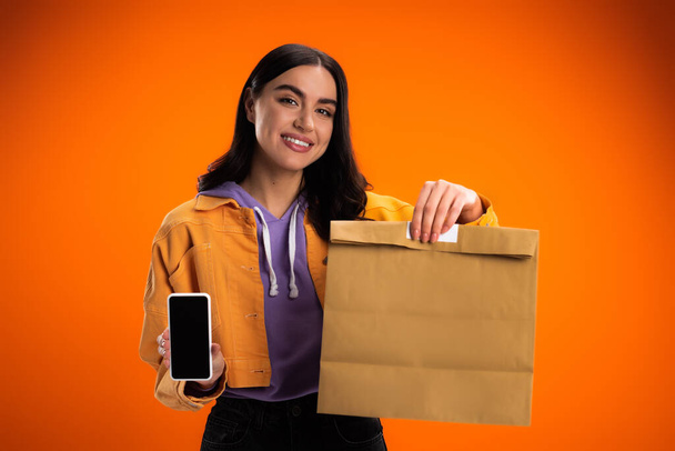 Positive young woman showing smartphone and paper bag isolated on orange - Foto, imagen