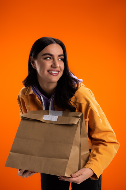 Young brunette woman holding paper bags and looking away isolated on orange - Foto, Imagen