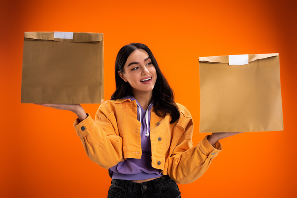 Позитивна молода жінка тримає паперові пакети з доставки їжі ізольовані на апельсині
 - Фото, зображення