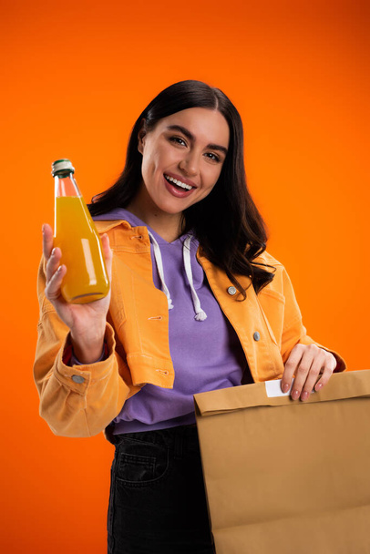 Positive young woman holding bottle with juice and paper bag isolated on orange - Photo, image