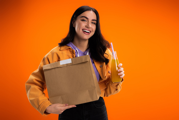 Cheerful young woman holding paper bag and juice in glass bottle isolated on orange - Valokuva, kuva